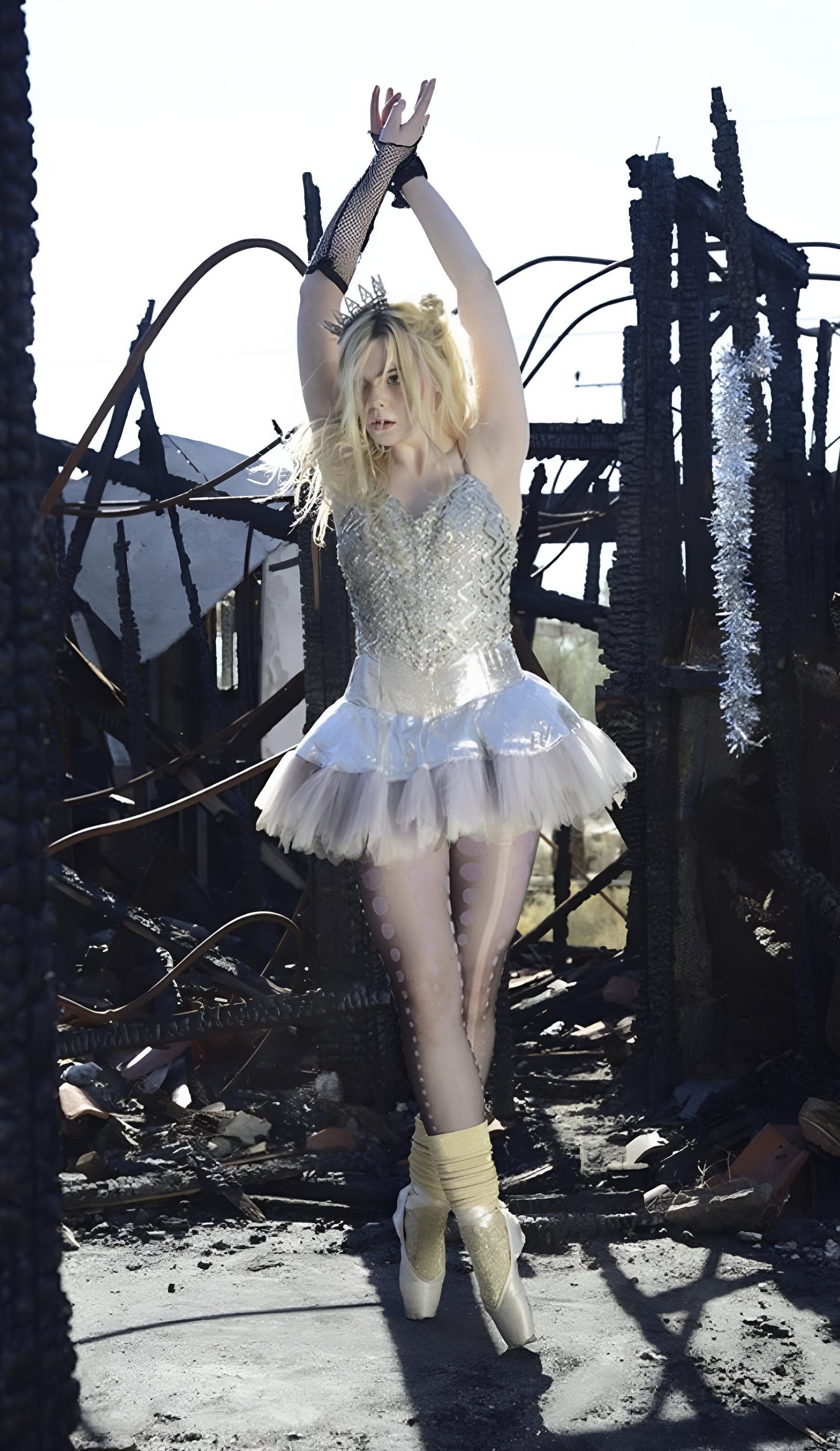 A ballerina in a white tutu poses with arms raised in front of a fire-damaged structure with a bright sky overhead in a full shot.