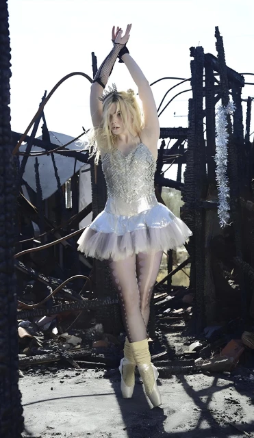 A ballerina in a white tutu poses with arms raised in front of a fire-damaged structure with a bright sky overhead in a full shot.