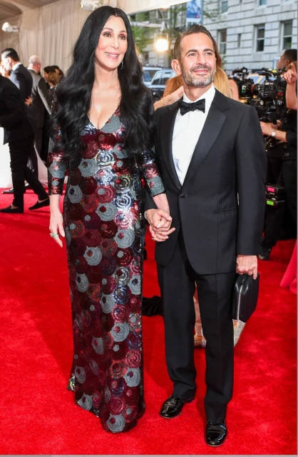 Cher and Marc Jacobs pose together on a red carpet. Cher wears a sparkling gown; Marc Jacobs is in a tuxedo.