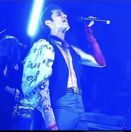 Male singer performing with microphone, wearing a vest and long gloves, with text on his sleeve, illuminated by blue stage lights.