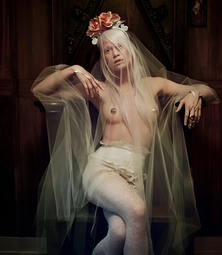 Surreal portrait of a woman in a sheer veil, wearing light-colored lingerie and sparkling tights, posing against a dark wooden backdrop.