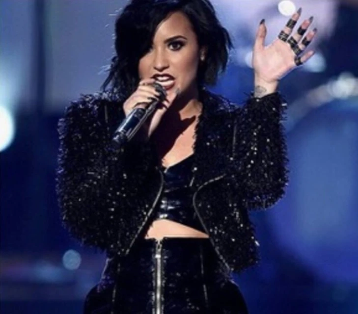 Adult female singer performing on stage with a microphone, wearing a black sparkly jacket and skirt, against a blue background.