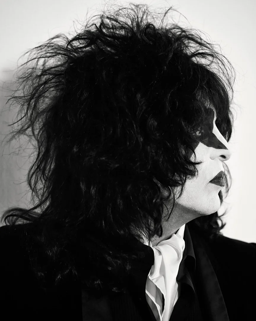 Monochrome portrait of a performer in theatrical makeup and long black hair, wearing a dark suit and white cravat against a white background.