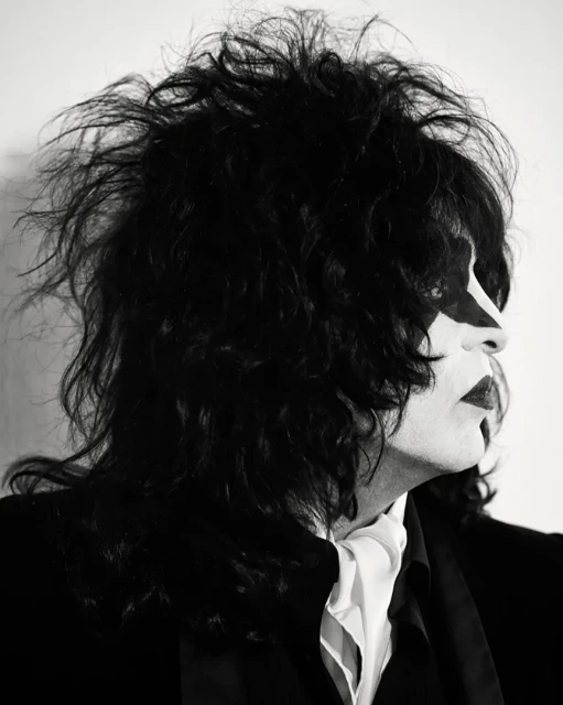 Monochrome portrait of a performer in theatrical makeup and long black hair, wearing a dark suit and white cravat against a white background.