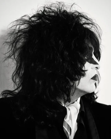 Monochrome portrait of a performer in theatrical makeup and long black hair, wearing a dark suit and white cravat against a white background.