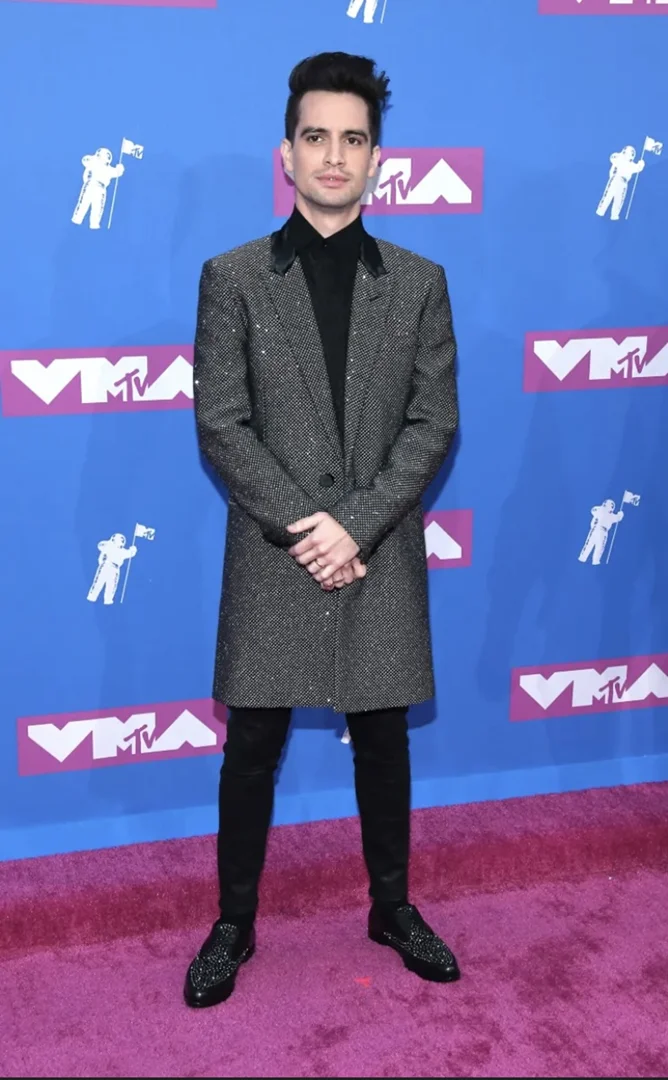 Brendon Urie on a red carpet wearing a sparkly coat, black pants, and black shoes standing in front of a VMA backdrop.