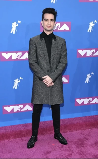 Brendon Urie on a red carpet wearing a sparkly coat, black pants, and black shoes standing in front of a VMA backdrop.
