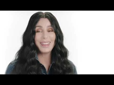 Close-up of Cher smiling in a denim jacket against a white background, showcasing her dark hair and makeup.