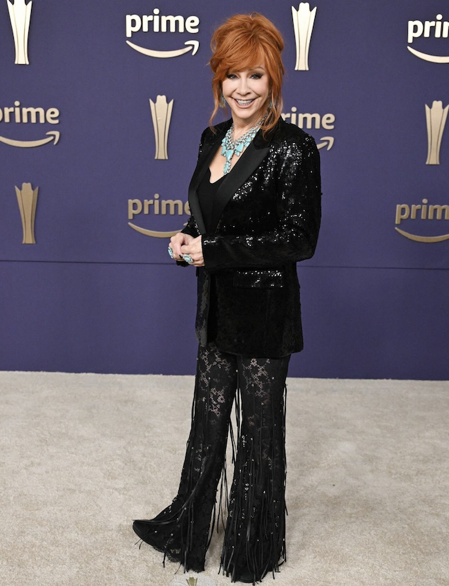 Reba McEntire poses on a carpet in a black sequin blazer, black lace fringe pants and turquoise jewerly in front of a Prime banner.