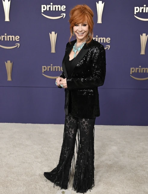 Reba McEntire poses on a carpet in a black sequin blazer, black lace fringe pants and turquoise jewerly in front of a Prime banner.