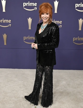 Reba McEntire poses on a carpet in a black sequin blazer, black lace fringe pants and turquoise jewerly in front of a Prime banner.