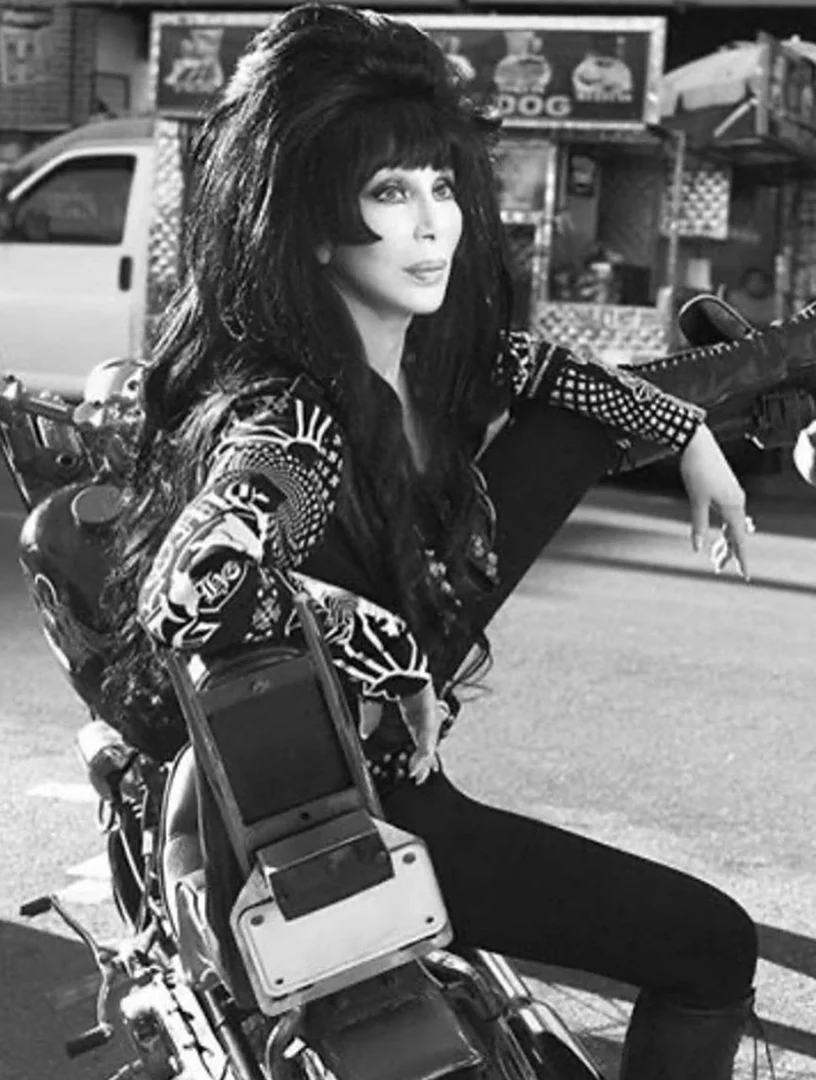 Cher, an adult woman with long dark hair, sits on a motorcycle in a black-and-white photo with a city street as background.