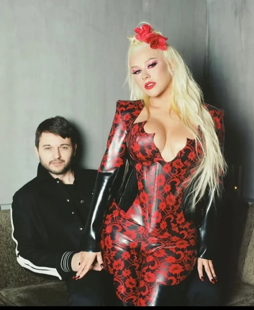 Christina Aguilera poses with a man in a black jacket. Aguilera wears a red and black floral latex suit, long blonde hair, and red lipstick.
