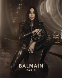 Cher wearing black latex catsuit and holding a black handbag with Balmain branding in a moody studio setting