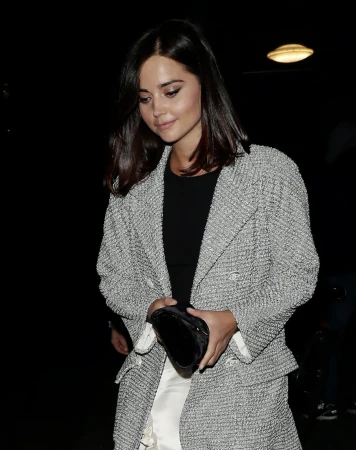 Jenna Coleman poses in a grey tweed jacket with a black top and white pants, holding a black clutch against a dark background.