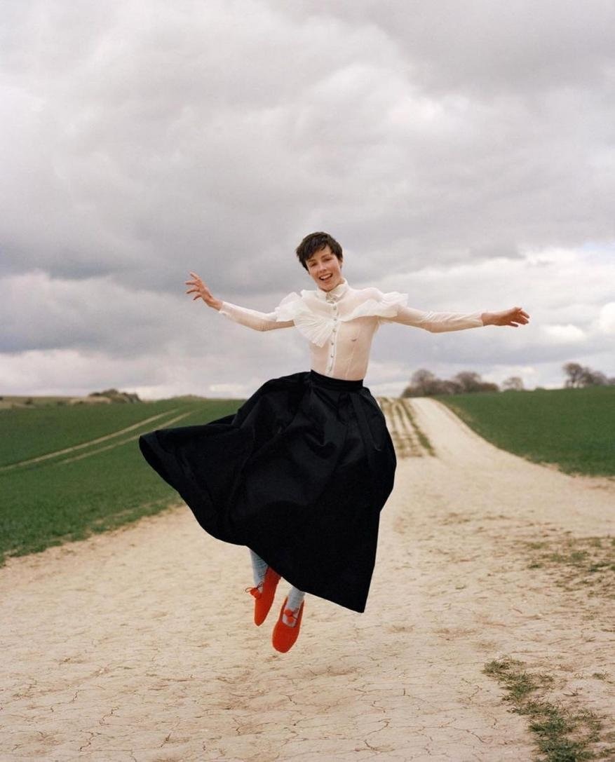 A cheerful woman in a white blouse and black skirt jumps on a dirt road lined with green fields under a cloudy sky, smiling widely.