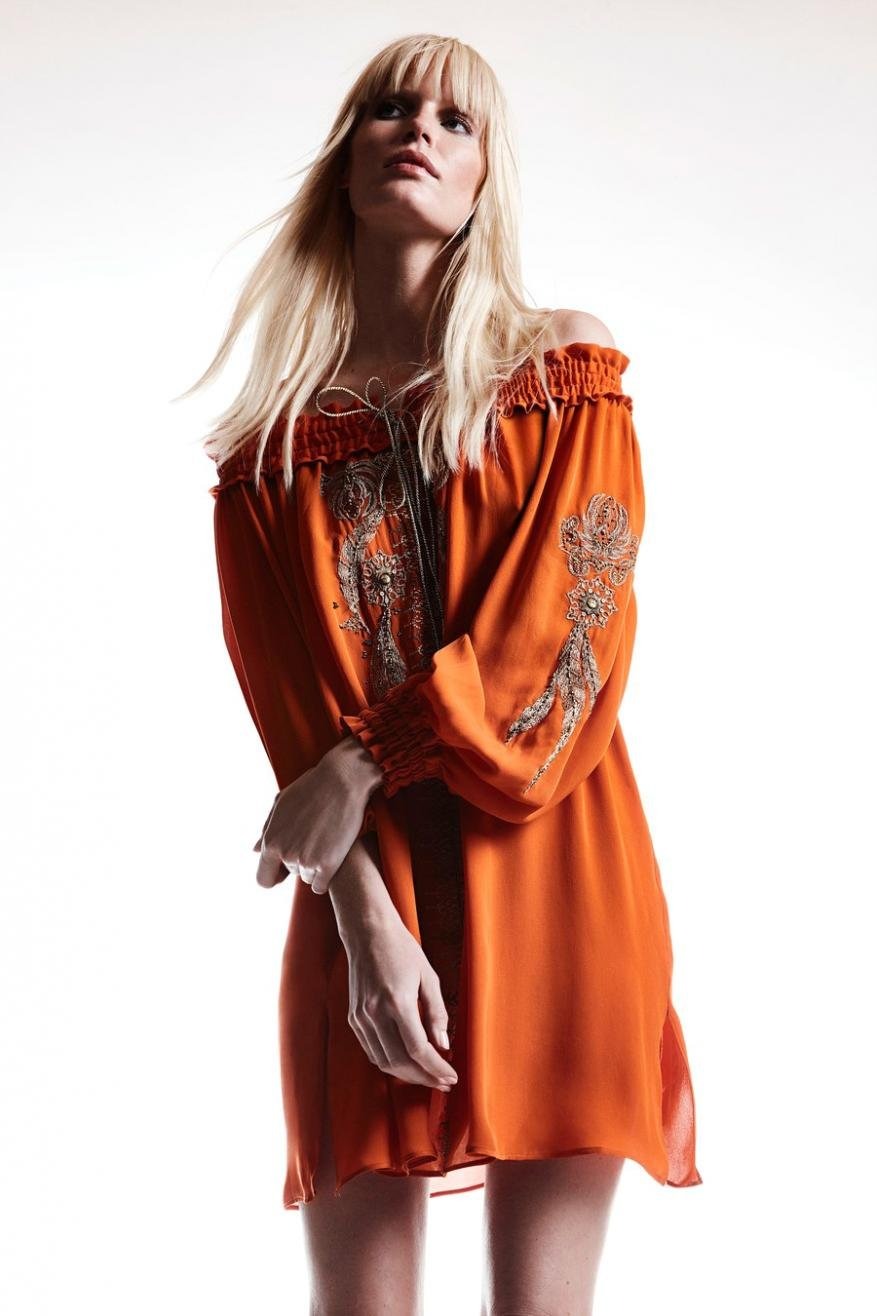 A young adult woman with blonde hair poses in an off-the-shoulder orange dress with beaded embellishments against a white backdrop.
