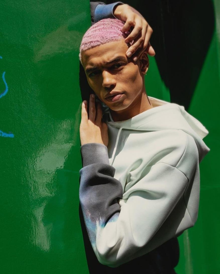 Portrait of a young adult man with pink hair, wearing a light-colored hoodie, leaning against a green wall in a close-up studio shot.