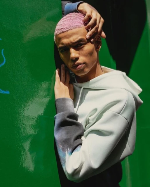 Portrait of a young adult man with pink hair, wearing a light-colored hoodie, leaning against a green wall in a close-up studio shot.