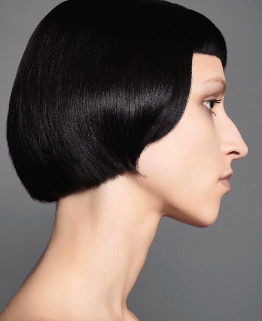 Profile view of a young adult woman with a short, blunt black bob haircut against a neutral gray backdrop in a beauty headshot.