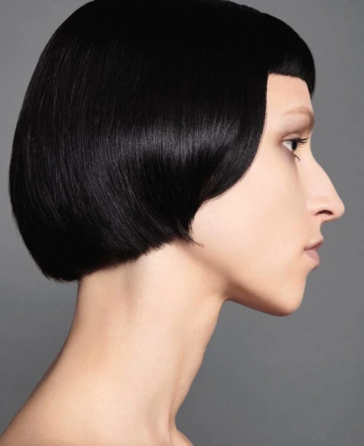 Profile view of a young adult woman with a short, blunt black bob haircut against a neutral gray backdrop in a beauty headshot.