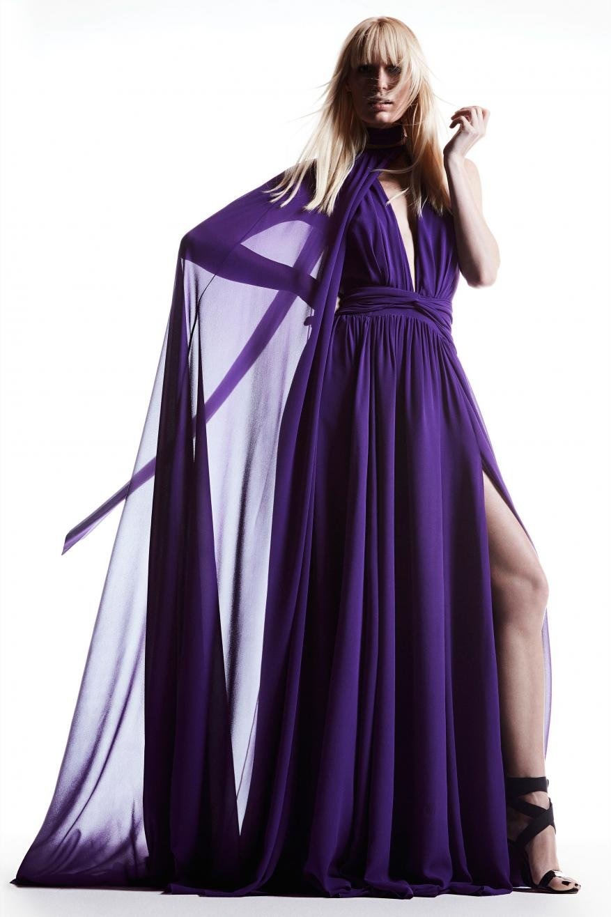 Fashion model in a flowing purple gown with a high slit and cape, posing in a photo studio with a white background and dramatic lighting.