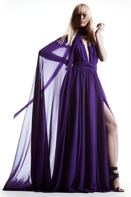 Fashion model in a flowing purple gown with a high slit and cape, posing in a photo studio with a white background and dramatic lighting.
