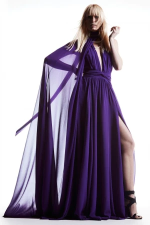 Fashion model in a flowing purple gown with a high slit and cape, posing in a photo studio with a white background and dramatic lighting.