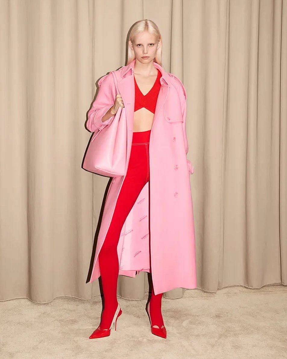 A fair-skinned adult female model poses in a pink trench coat, red top, leggings, and high heels in front of a tan curtain backdrop.