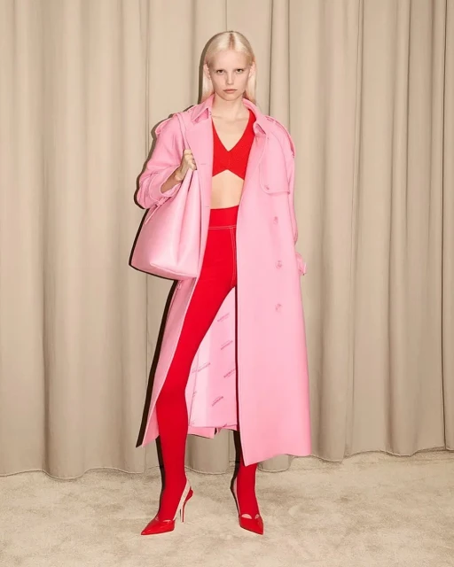 A fair-skinned adult female model poses in a pink trench coat, red top, leggings, and high heels in front of a tan curtain backdrop.