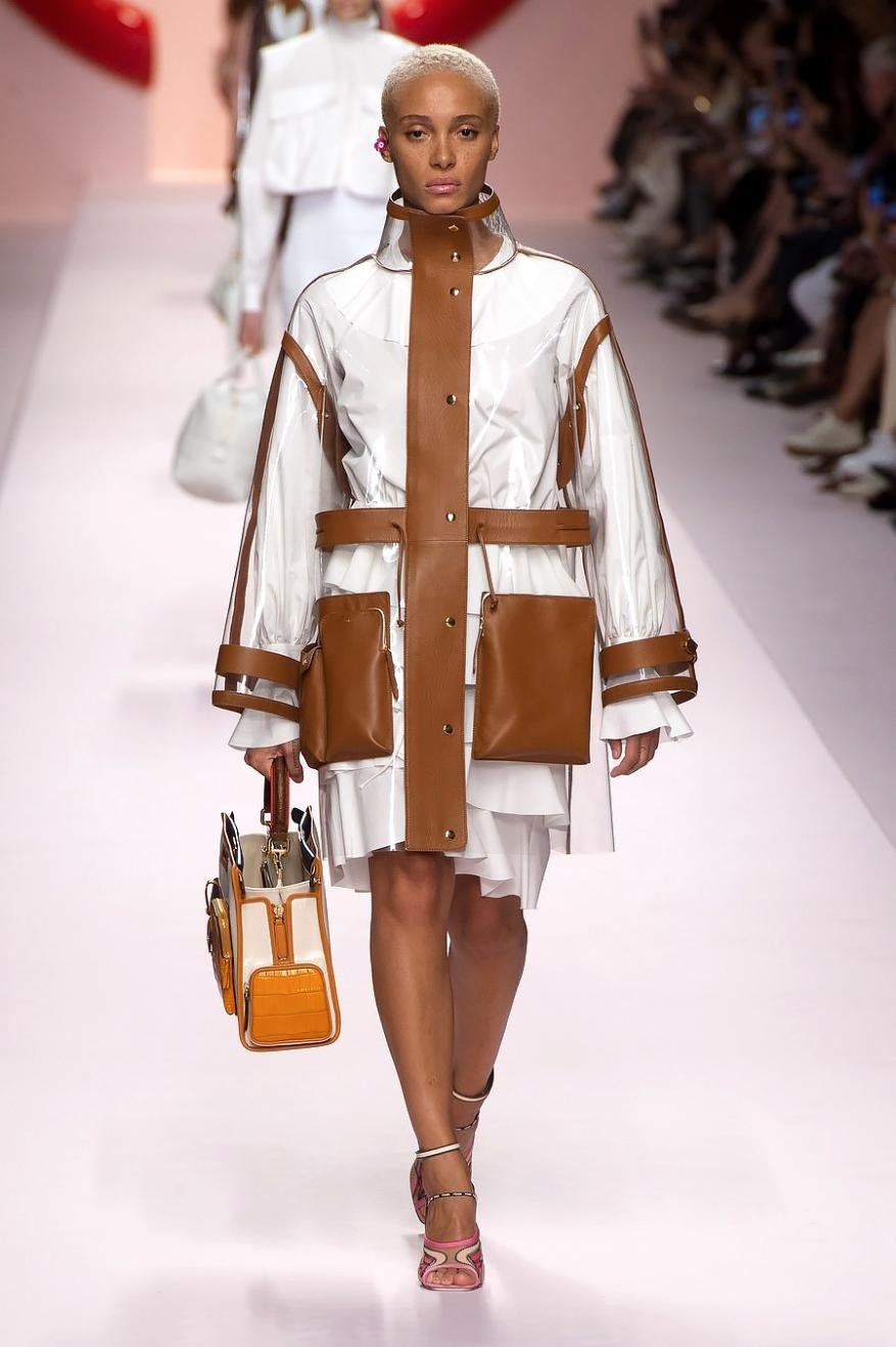 Runway fashion: a woman models a white dress with a transparent jacket accented with brown pockets and trim, carrying a matching handbag.