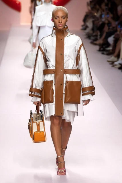 Runway fashion: a woman models a white dress with a transparent jacket accented with brown pockets and trim, carrying a matching handbag.