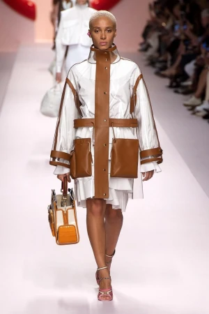 Runway fashion: a woman models a white dress with a transparent jacket accented with brown pockets and trim, carrying a matching handbag.