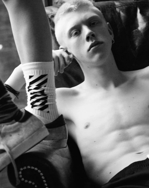 Black and white portrait of a young adult male with blond hair, bare chest, earring, with sock and athletic shoe visible.