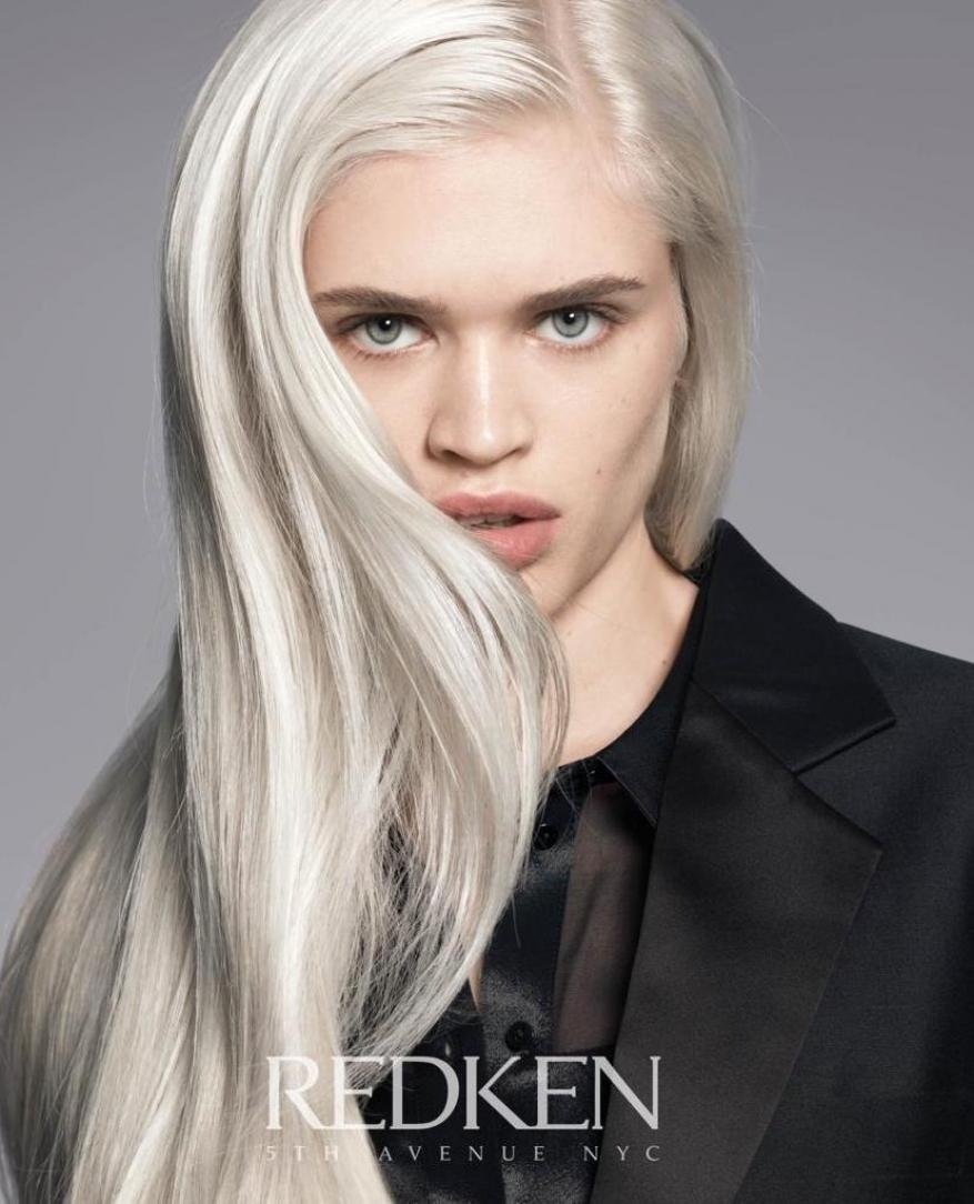Close-up shot of a model with long, straight, platinum blonde hair, wearing a black blazer and top, against a grey backdrop.