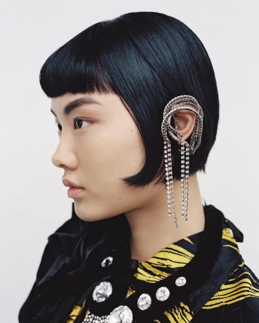 Close-up of a stylish woman with short black hair and distinctive earrings, set against a clean white background, showcasing fashion and beauty.
