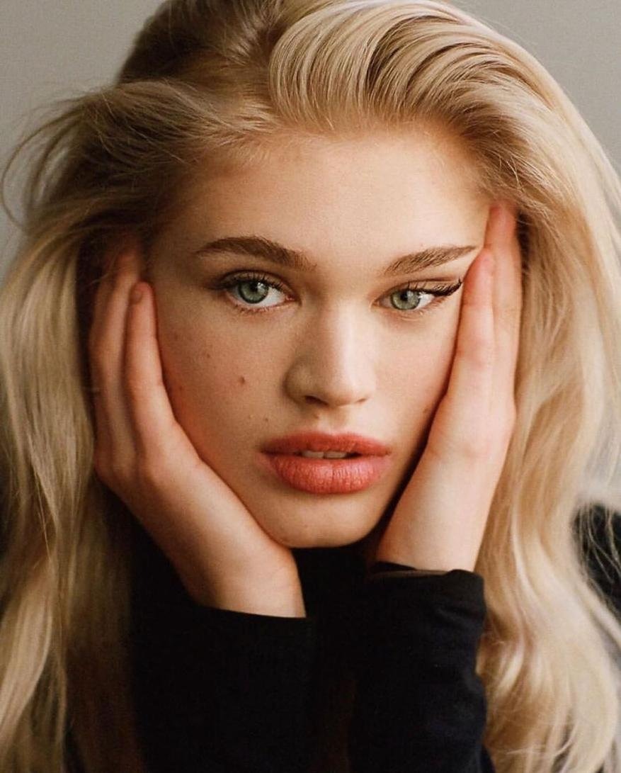 Close-up portrait of a young woman with long blonde hair holding her face in her hands, wearing a black top against a neutral background.