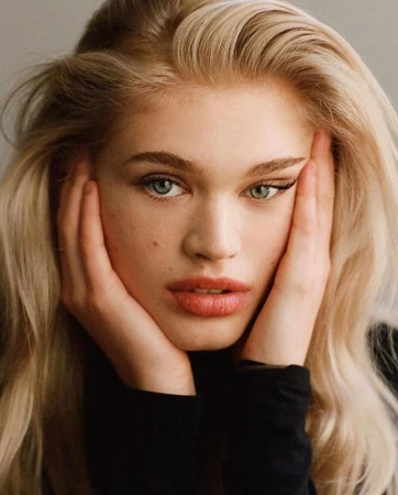 Close-up portrait of a young woman with long blonde hair holding her face in her hands, wearing a black top against a neutral background.