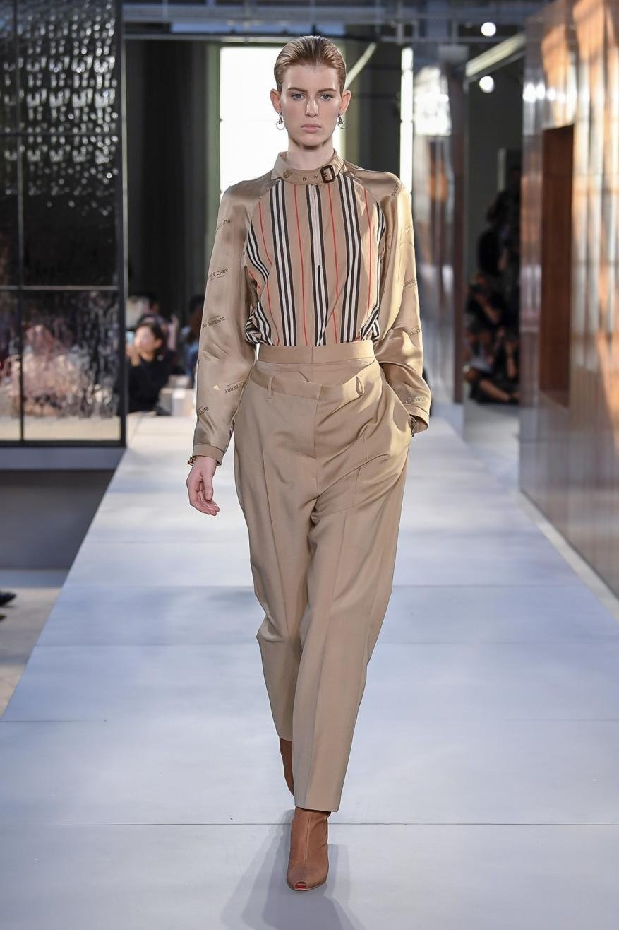 Runway fashion show featuring a female model in a beige striped blouse and trousers, accessorized with earring, walking on a light grey catwalk.
