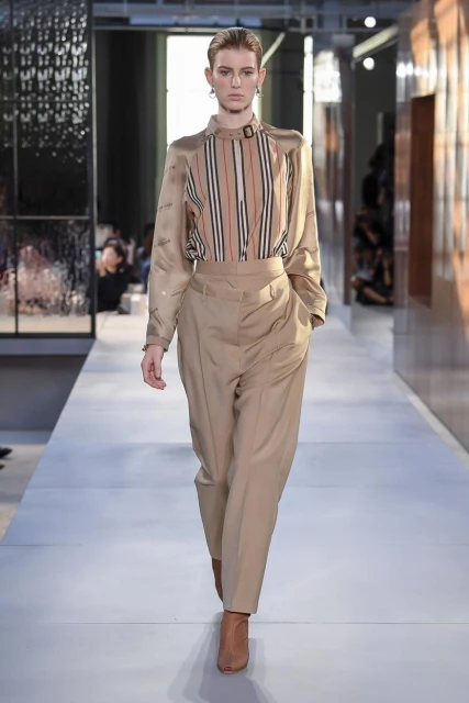 Runway fashion show featuring a female model in a beige striped blouse and trousers, accessorized with earring, walking on a light grey catwalk.