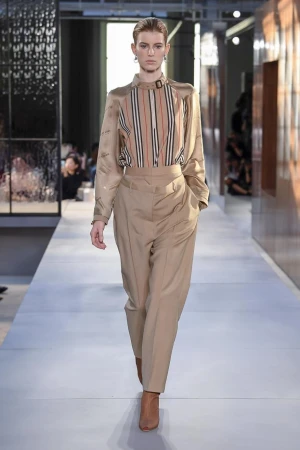 Runway fashion show featuring a female model in a beige striped blouse and trousers, accessorized with earring, walking on a light grey catwalk.