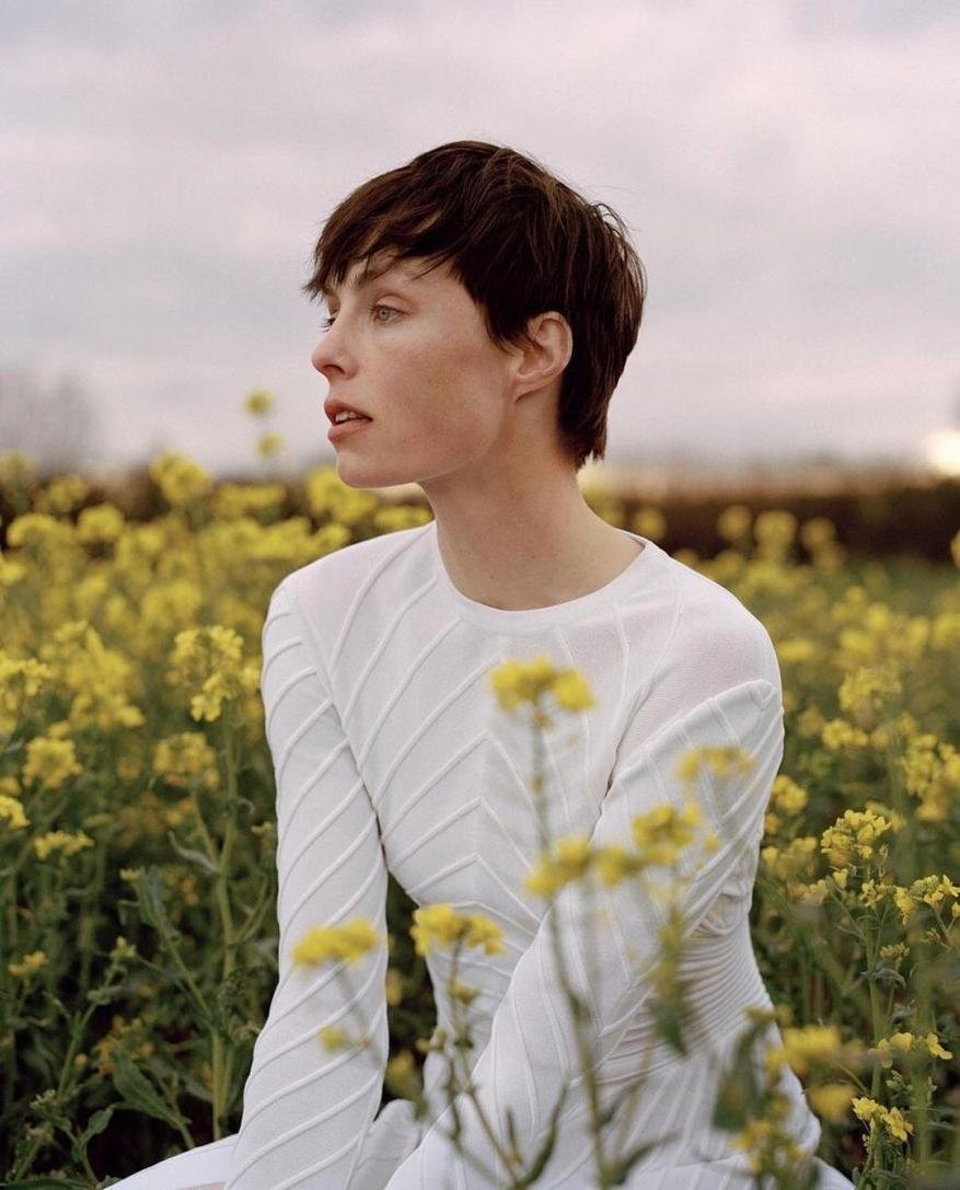 Woman with short brown hair poses in a field of yellow flowers wearing a white dress, looking off to the side with a neutral expression.