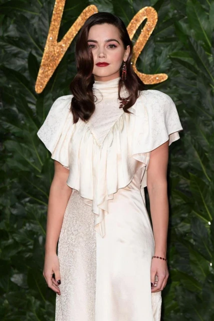 Portrait of a brunette woman with dark red lipstick wearing a cream-colored dress standing against a leafy green and brown backdrop.