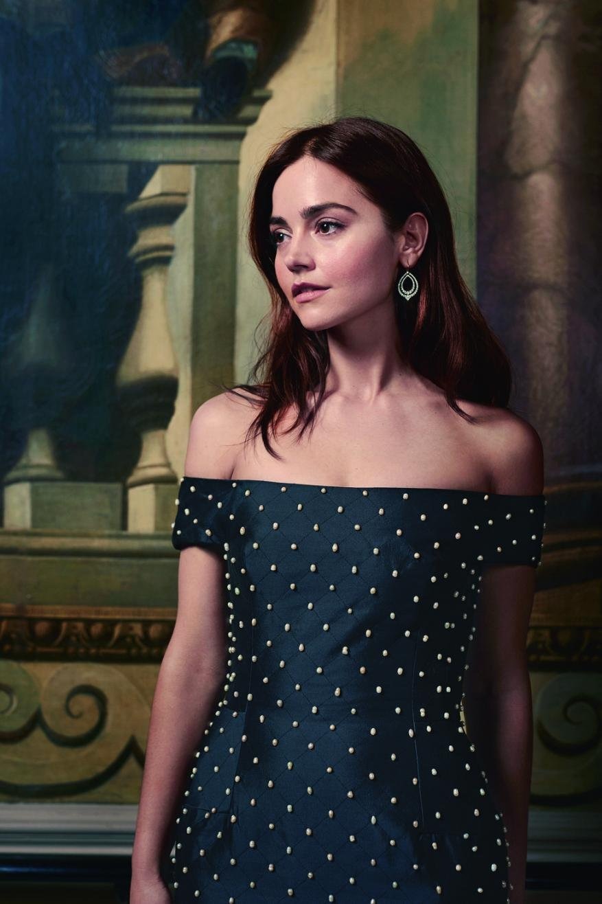 Portrait of a woman with dark hair wearing an off-the-shoulder dress with pearls, standing against a backdrop of an old painting.
