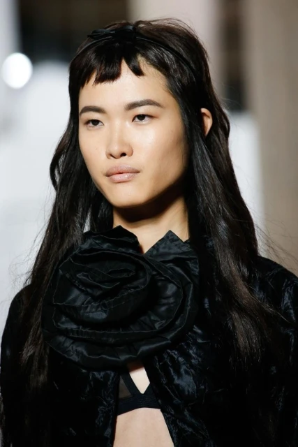 Portrait of an Asian female fashion model with bangs and long hair, wearing a black top with a flower detail at the neckline.
