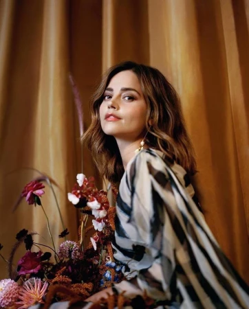 Portrait of a woman with layered brown hair wearing a patterned dress, posed among floral arrangements against a draped golden curtain background.