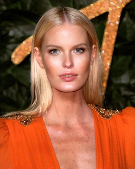 Blonde woman with fair skin and blue eyes poses in front of a leafy background, wearing an orange dress with shoulder embellishments.