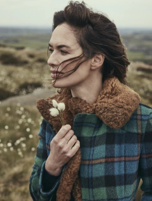 A woman in a plaid coat with a fur collar stands in an open, grassy field holding small flowers. Her hair is styled upwards.
