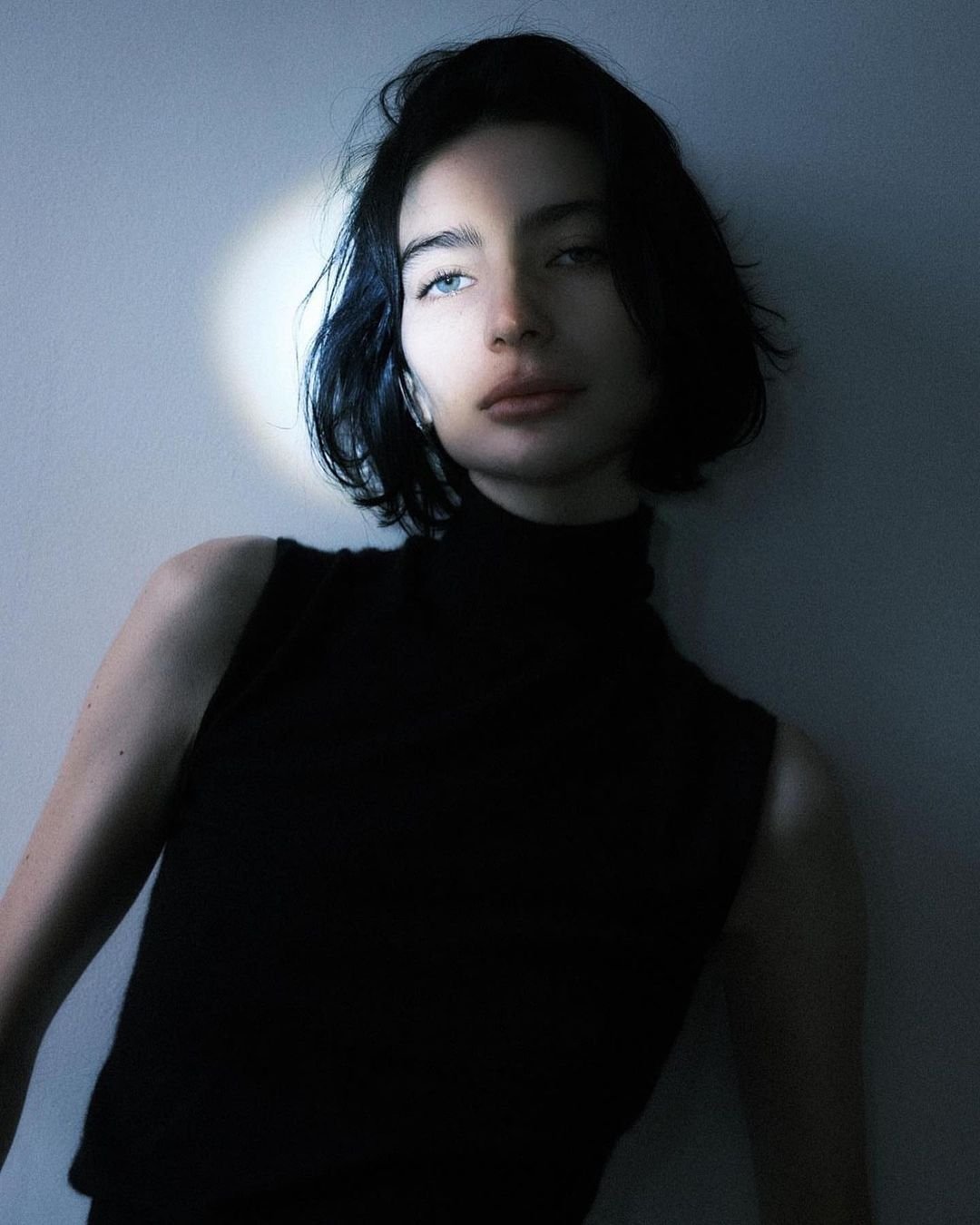 Portrait of a young woman with dark hair and a black top leaning against a light blue wall in soft, diffused studio lighting.