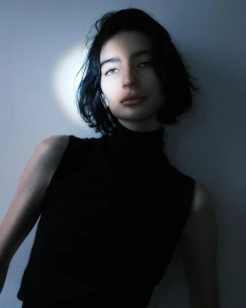 Portrait of a young woman with dark hair and a black top leaning against a light blue wall in soft, diffused studio lighting.